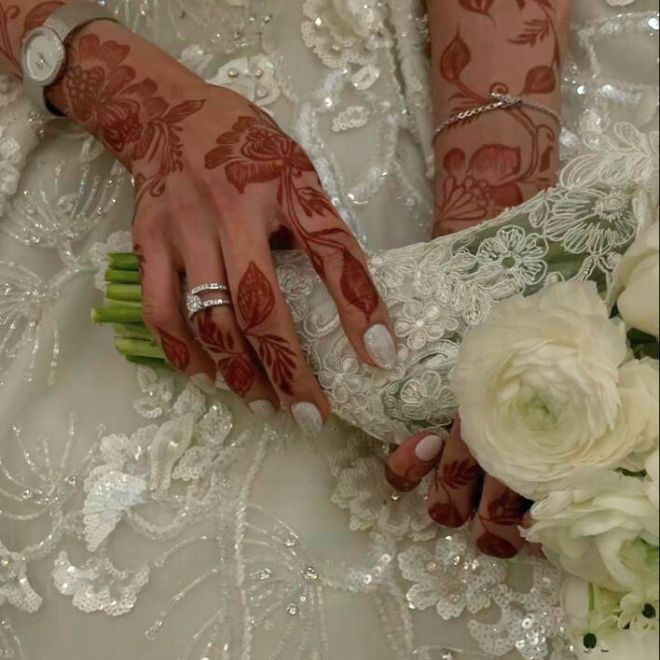 يد عروس خليجية مزينة بنقش حناء داكن تمسك باقة ورد
بيضاء فوق فستان زفاف مطرز