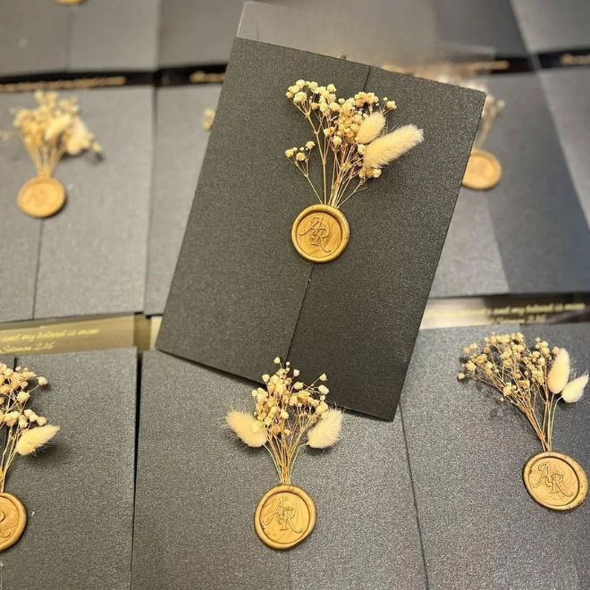 بطاقة دعوة زفاف فاخرة باللون الأسود مع ختم شمع ذهبي
وزهور مجففة تمثل تجهيز دعوات حفل الزواج وإرسالها للمدعوين في
السعودية